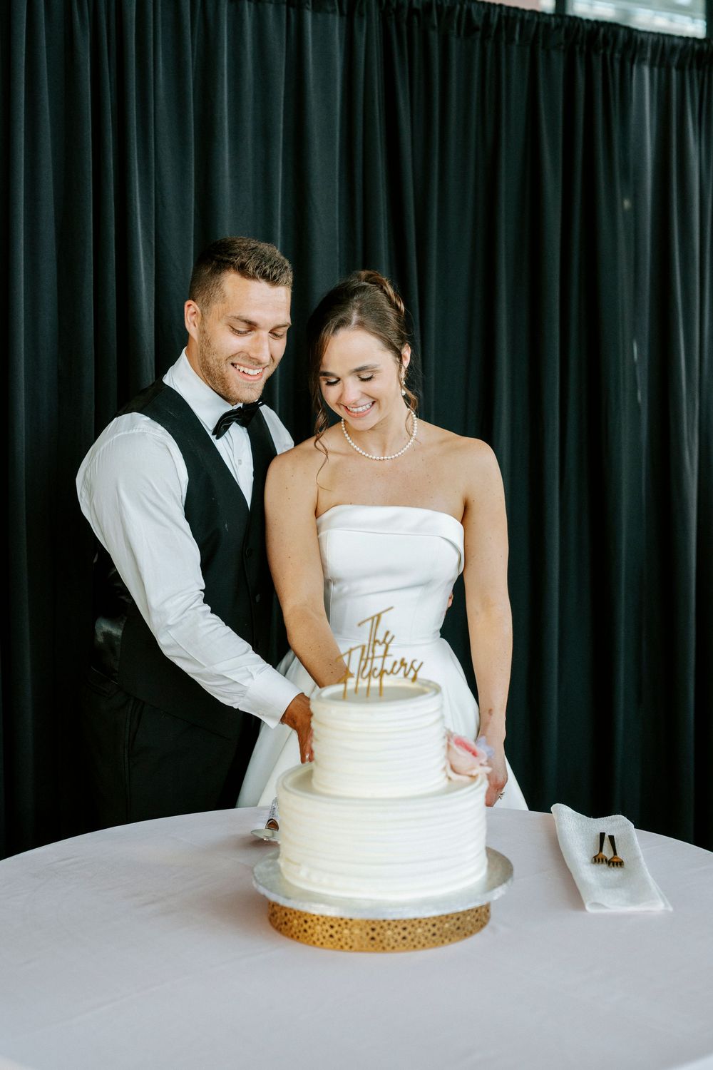 Bride and groom smiling while cutting their white three-tier wedding cake together at their reception.