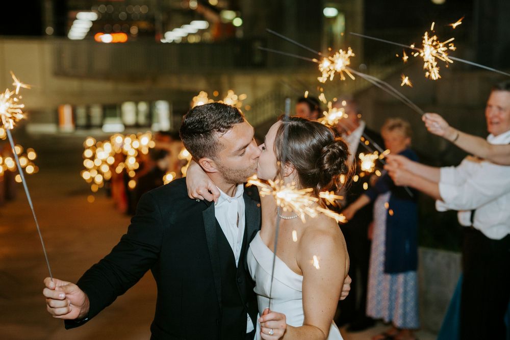 Bride and groom share a kiss surrounded by guests holding sparklers at their wedding reception.
