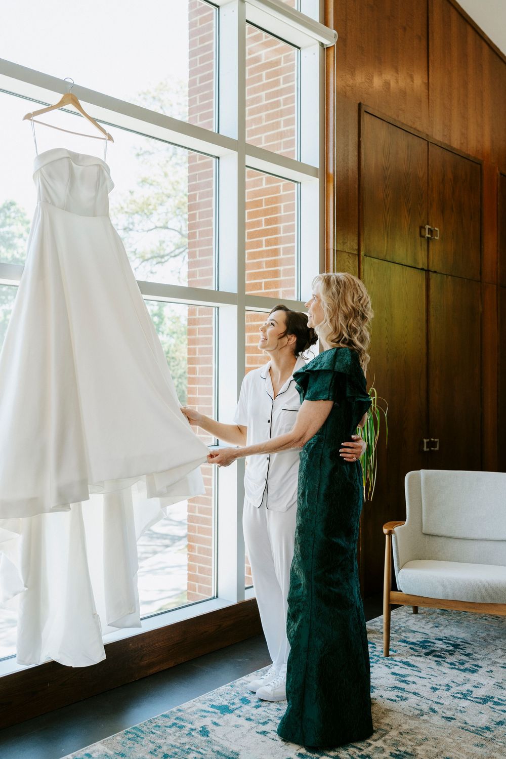 Bride and bridesmaid admiring a white wedding gown hanging by a large window in an elegant room.
