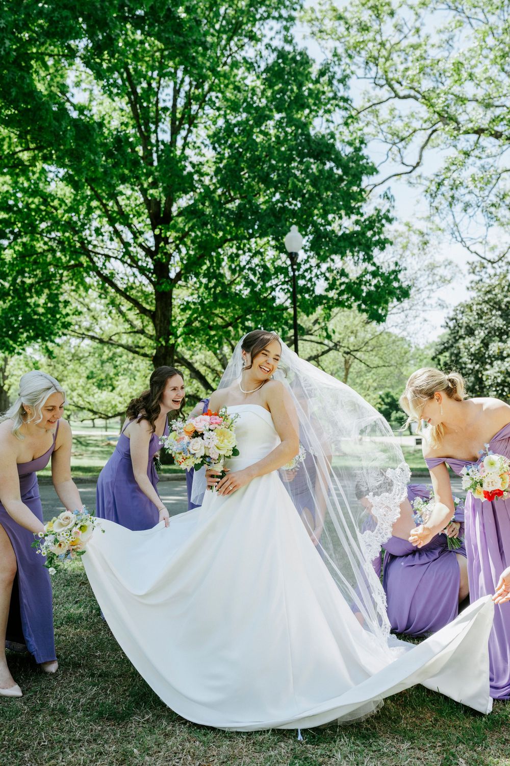 Bridesmaids in purple dresses help bride with flowing white veil and gown in a lush green park.