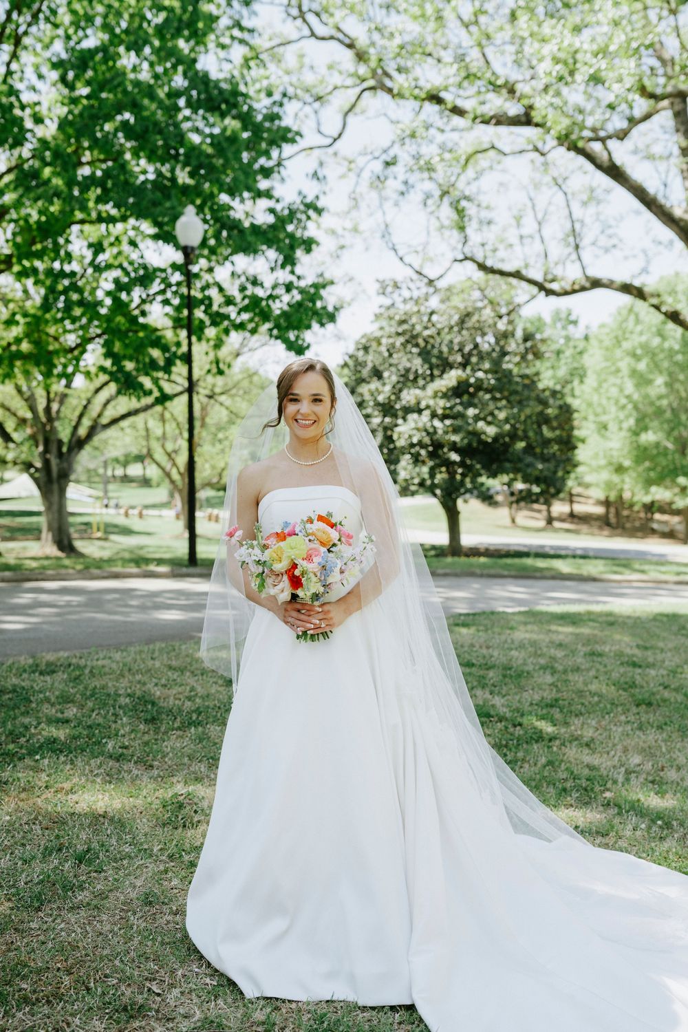 Smiling bride in a strapless white gown and veil holds a colorful bouquet in a sunny park.