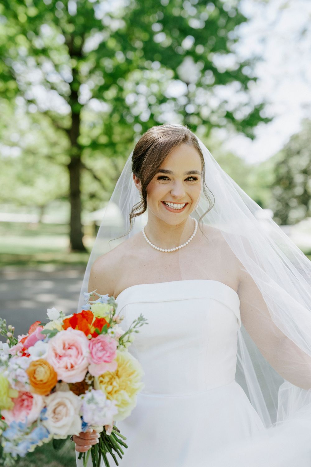 Smiling bride in strapless white gown and veil holds colorful bouquet outdoors among lush green trees.