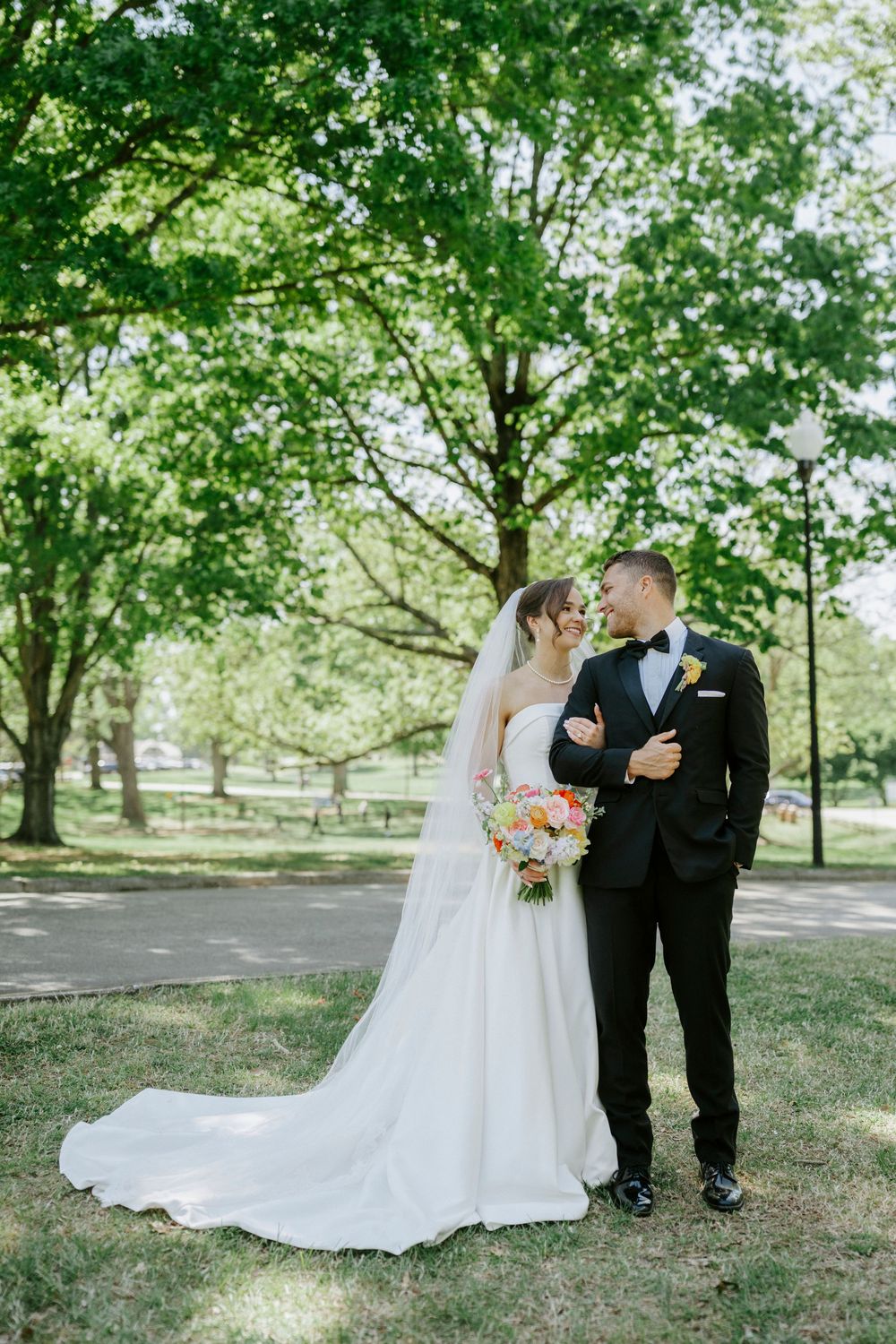 Bride and groom share a tender moment under lush green trees in a sunlit park setting.