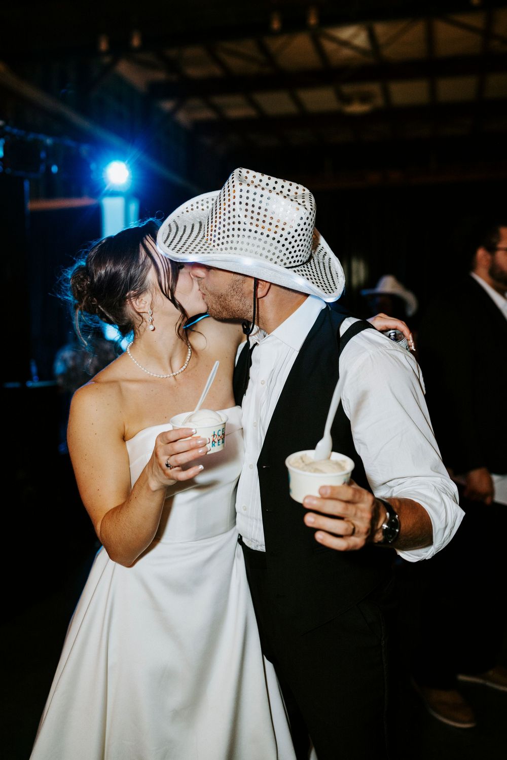 Bride and groom kiss on the dance floor, groom wearing a sequin cowboy hat at a lively wedding reception.