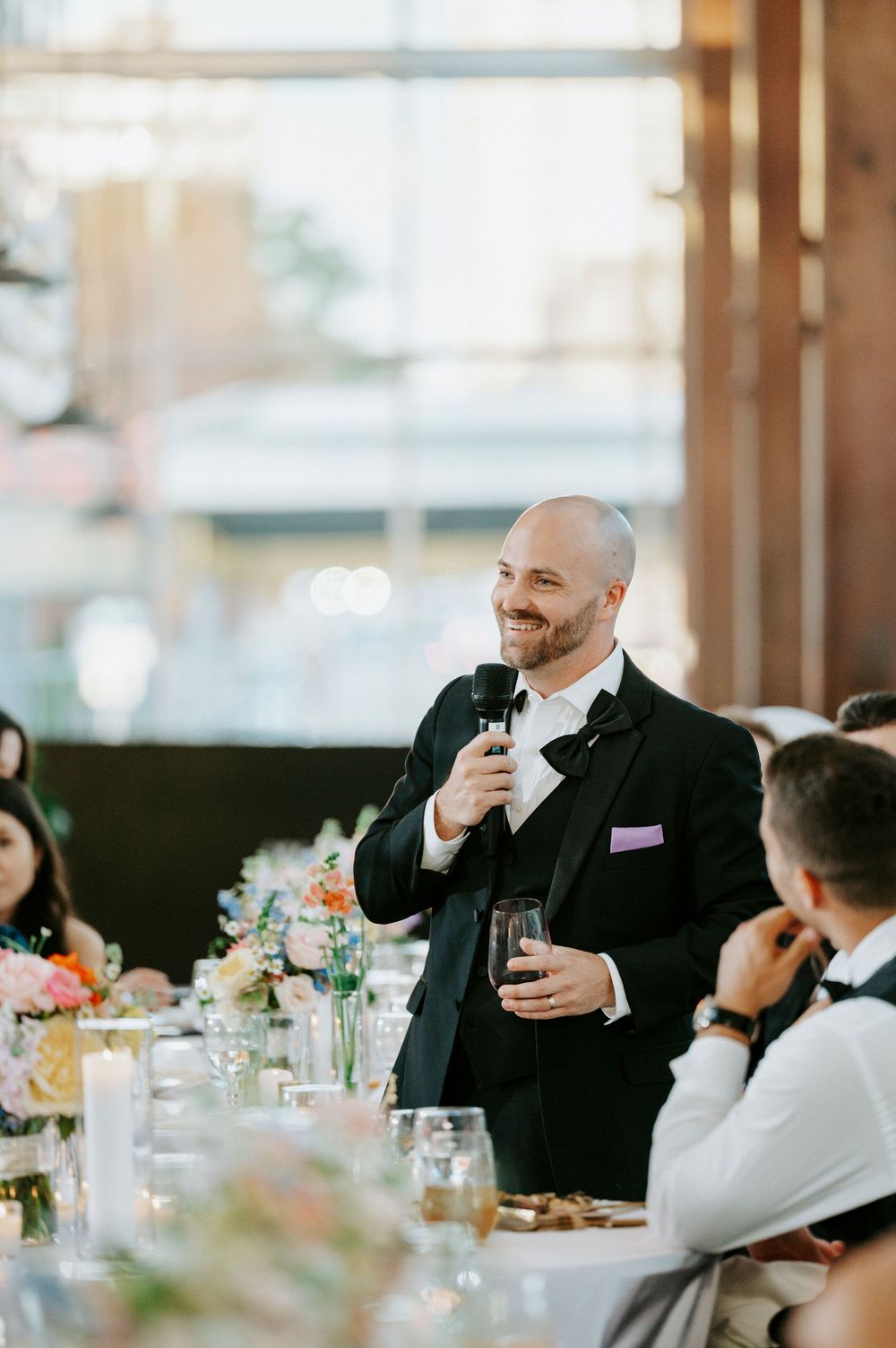 Smiling bald groom in a tuxedo gives a wedding toast holding a microphone and drink glass.