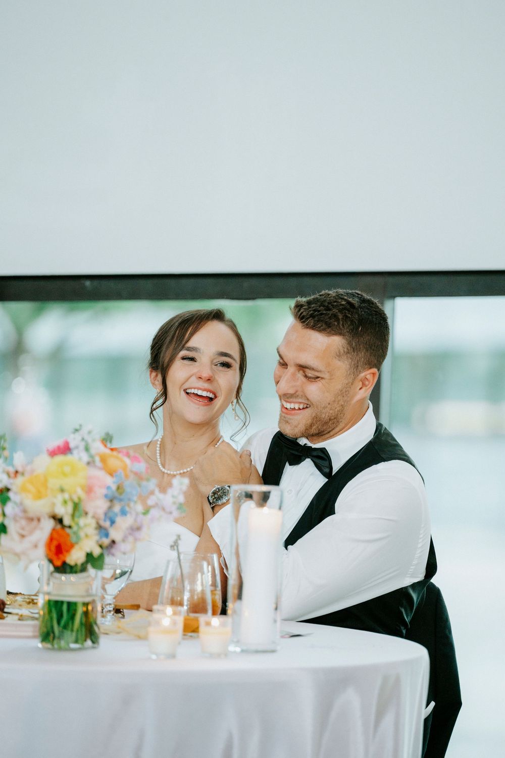 Bride and groom laugh joyfully at sweetheart table with floral centerpiece and candles at wedding reception.