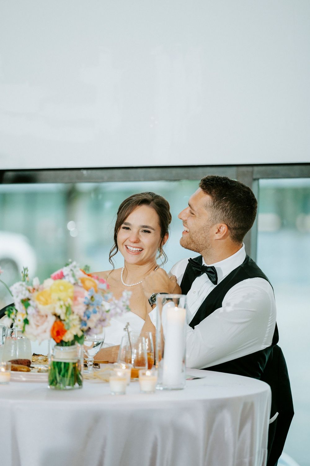 Smiling bride and groom seated at sweetheart table with colorful floral centerpiece during wedding reception.