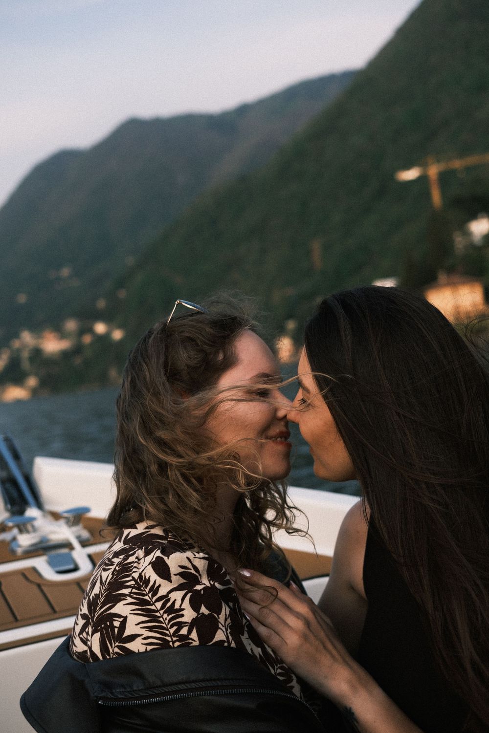 Two women sharing a romantic kiss on a boat with scenic mountain lake views in the background.