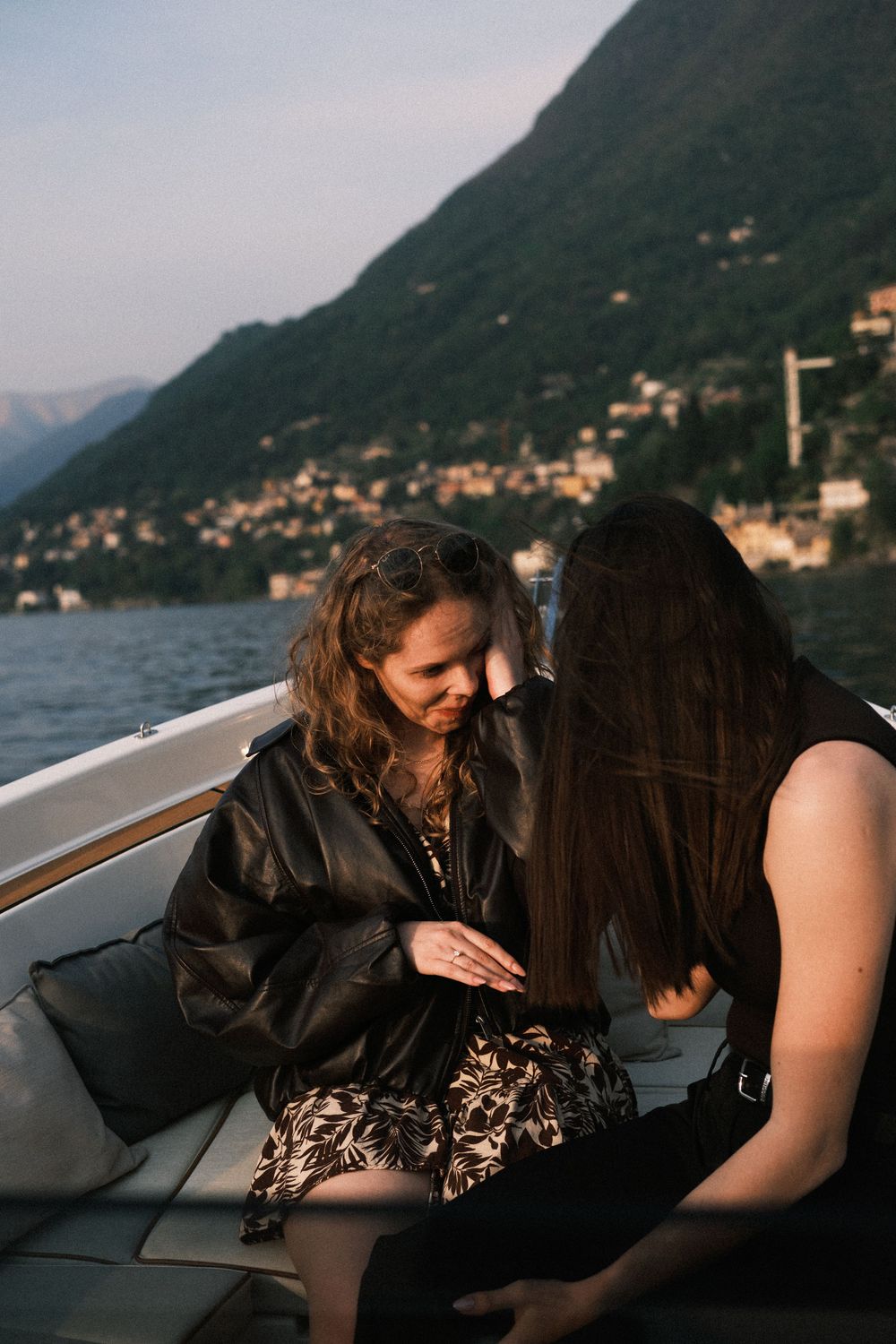 Two women share an intimate moment on a boat with scenic lake and mountain village backdrop at dusk.