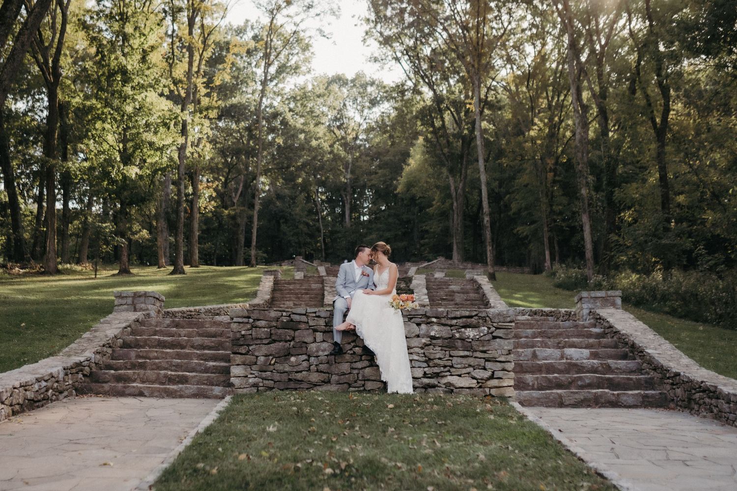 Wedding couple sitting on stone steps surrounded by lush green trees in an outdoor garden ceremony setting at Percy Warner State Park.