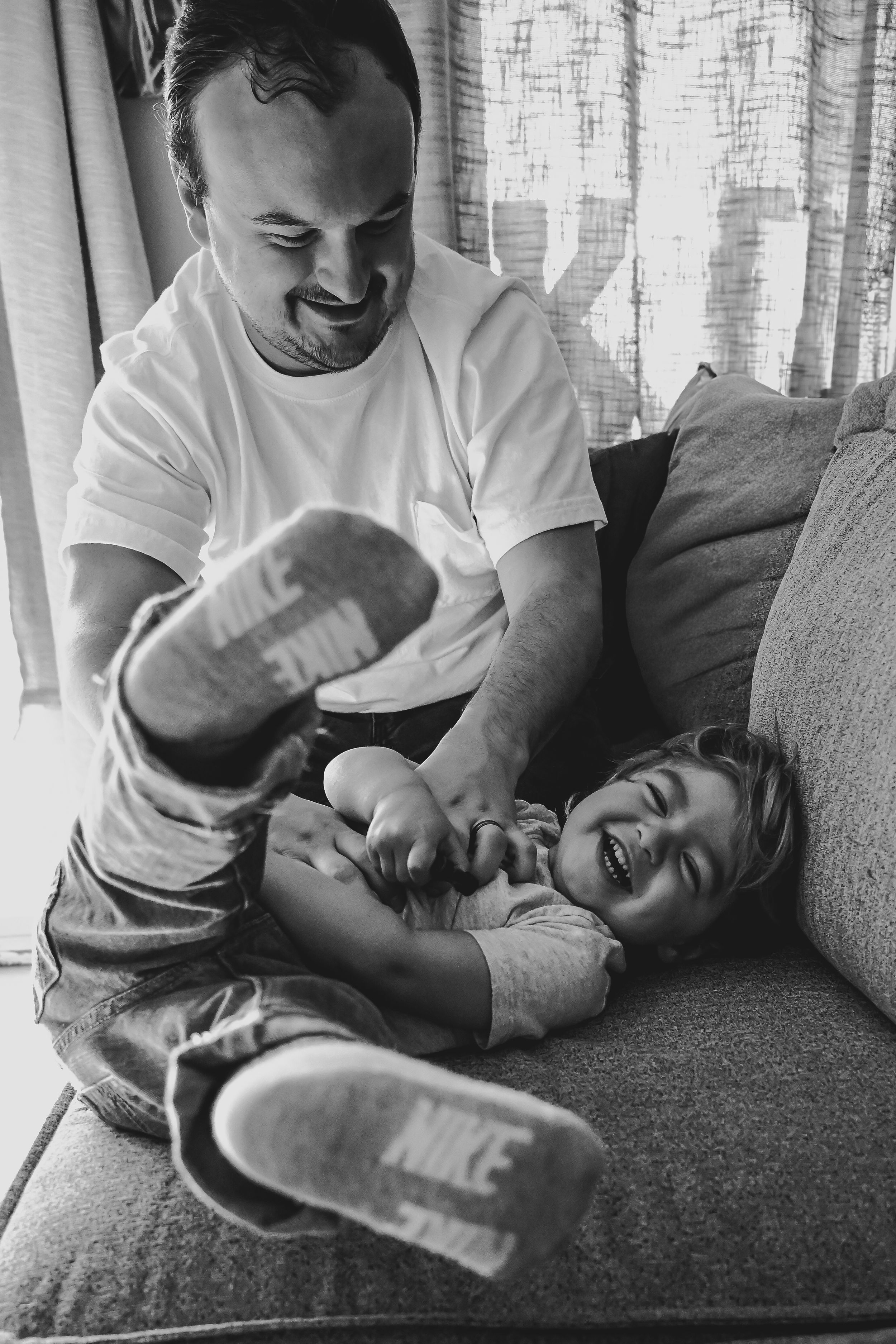 Black and white photo of a father tickling his laughing child on a couch, both wearing Nike shoes.