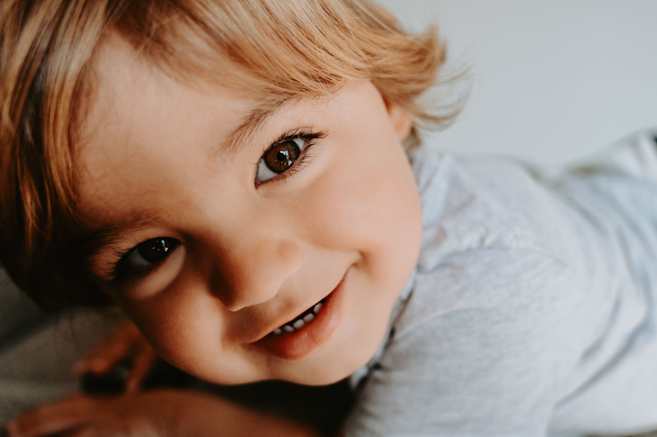 Smiling toddler with blonde hair and bright eyes looking at camera in a close-up portrait photo.