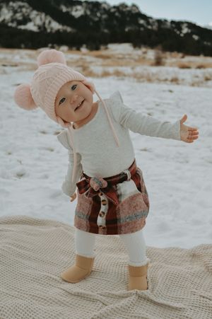 Adorable baby in pink pom-pom hat, plaid skirt, and tan boots stands in snowy field with arms outstretched.