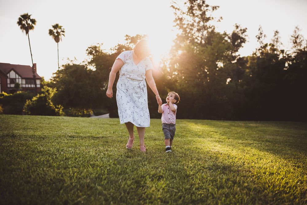 Inland Empire mother and son walking together in a grassy field at sunset with golden sunlight streaming through the trees.