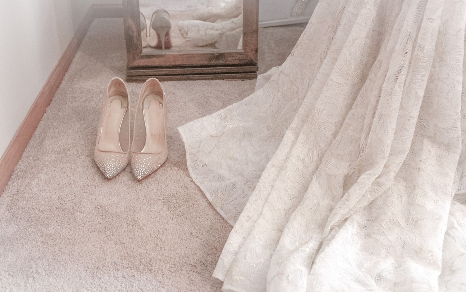 A pair of glittery silver high heel shoes with red soles sits on beige carpet next to wooden furniture.