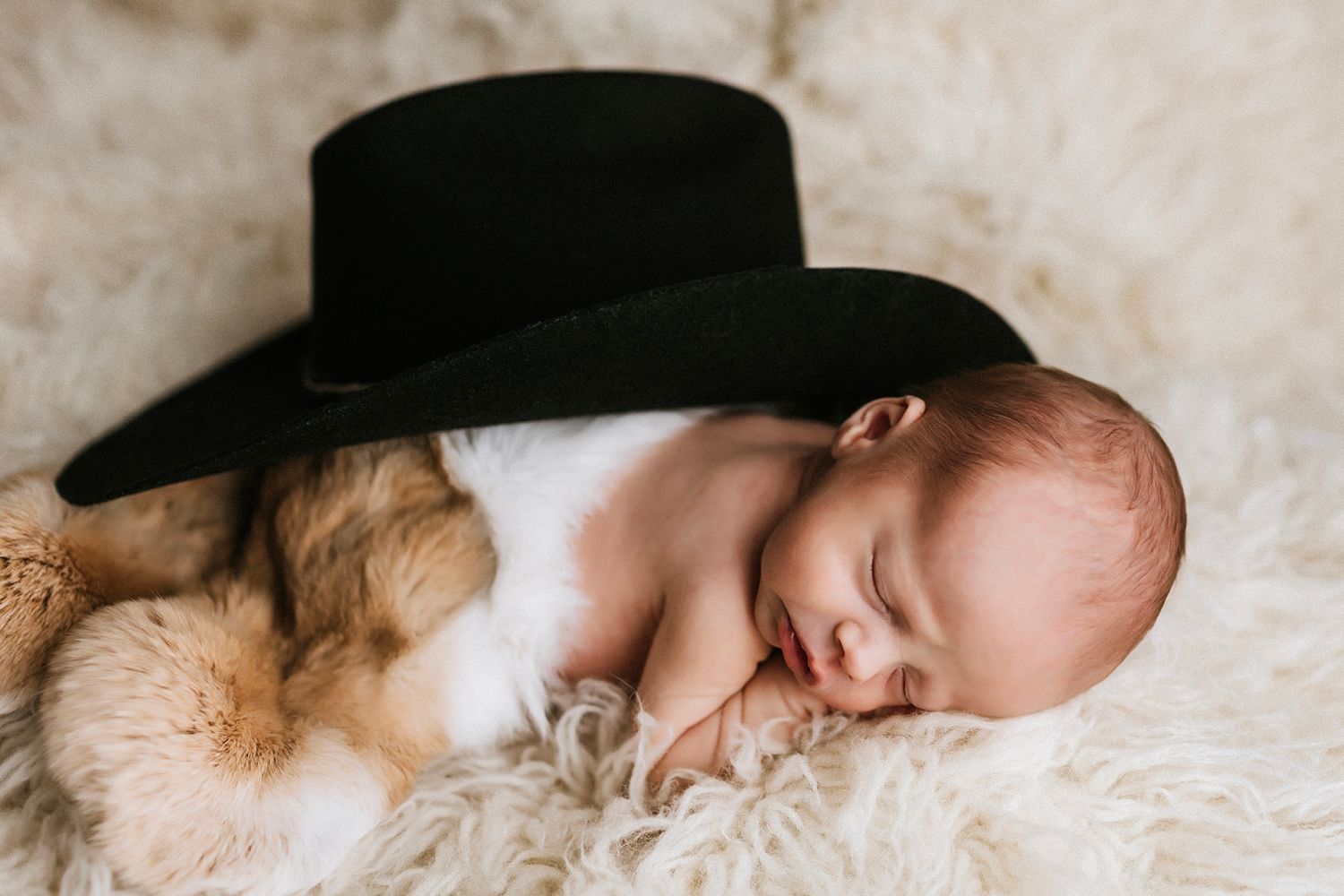 Sleeping newborn wearing black hat rests peacefully on soft white fur blanket.
