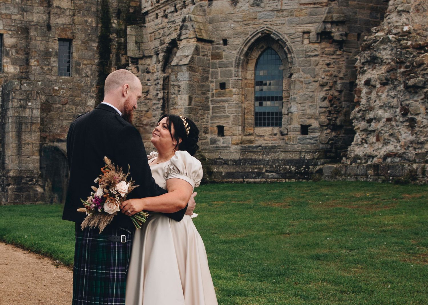 Wedding portrait in front of historic stone castle ruins with Scottish kilt attire.