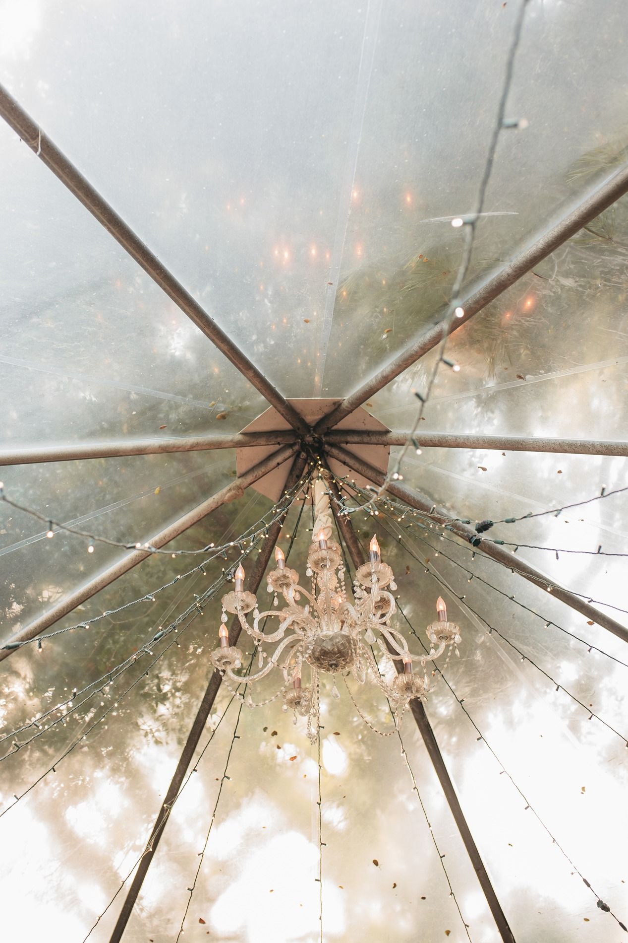 Looking up at an elegant crystal chandelier through a transparent tent canopy with water droplets and soft glowing lights.