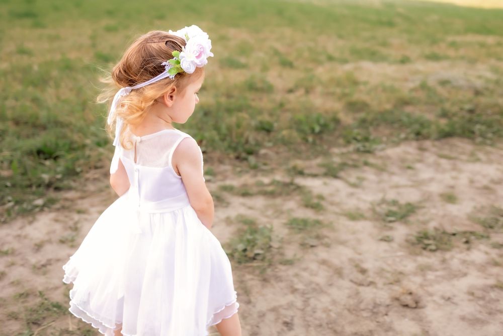 A child in a white dress twirls and dances outdoors in a field during a sunny summer day.