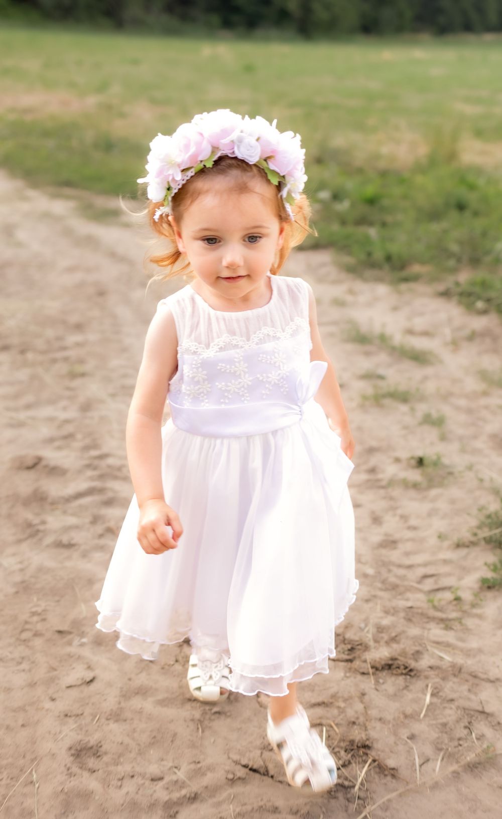 A child in a white lacy dress and floral crown walks along a dirt path in a sunny outdoor setting.