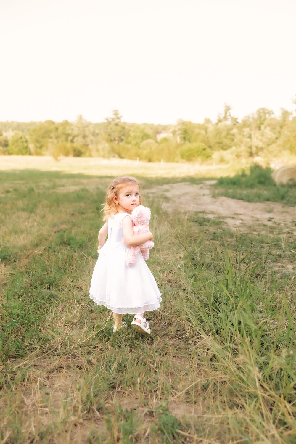 A small child wearing a white dress walks through a grassy field at sunset.