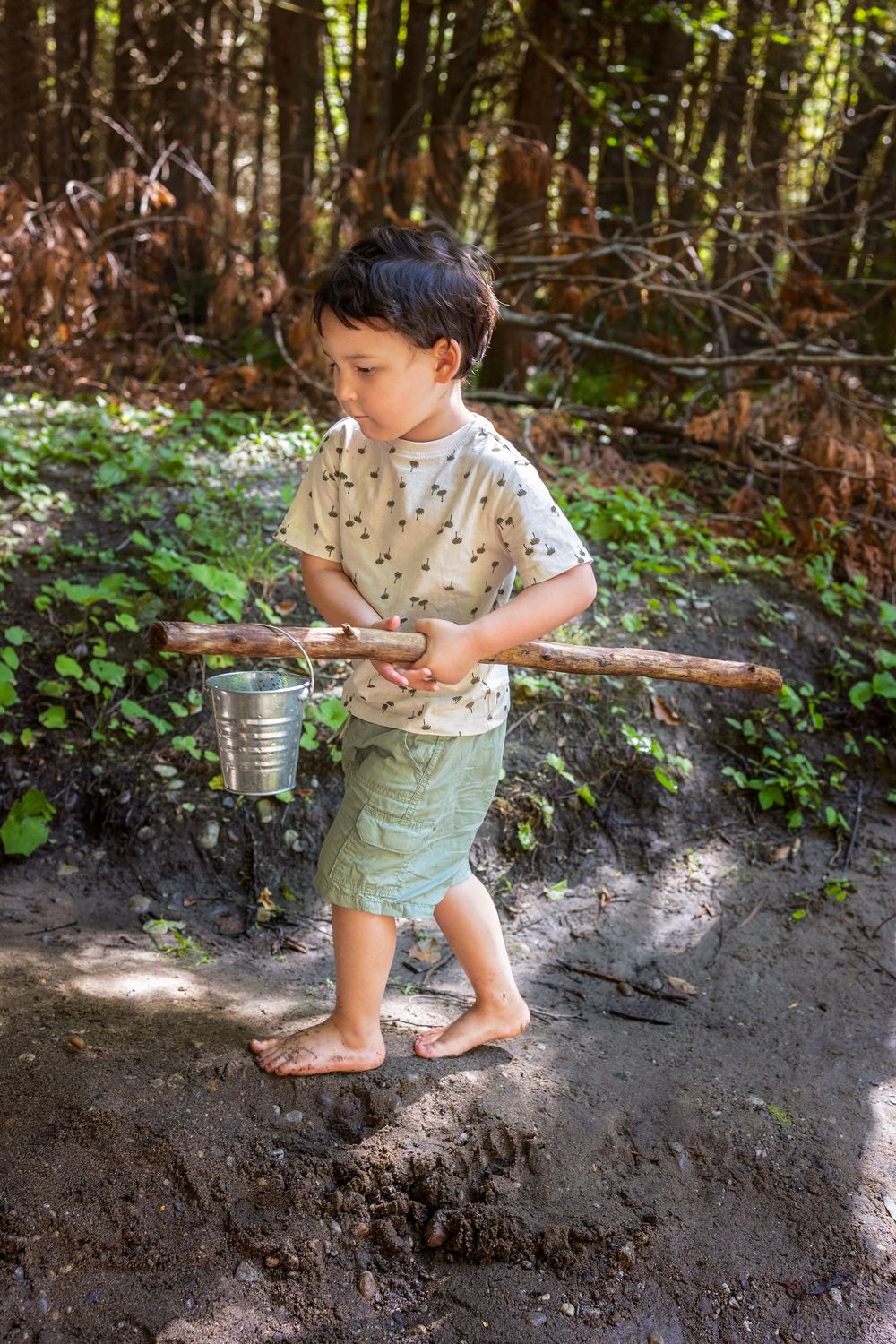 A child plays in a sunlit forest clearing while carrying a metal pail and wearing summer clothing.