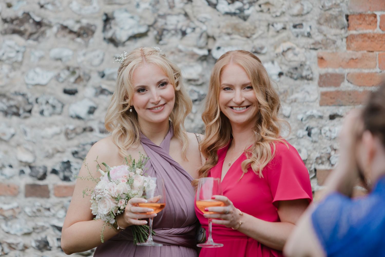 Two people in formal attire smile and toast with wine glasses against a rustic stone wall background.
