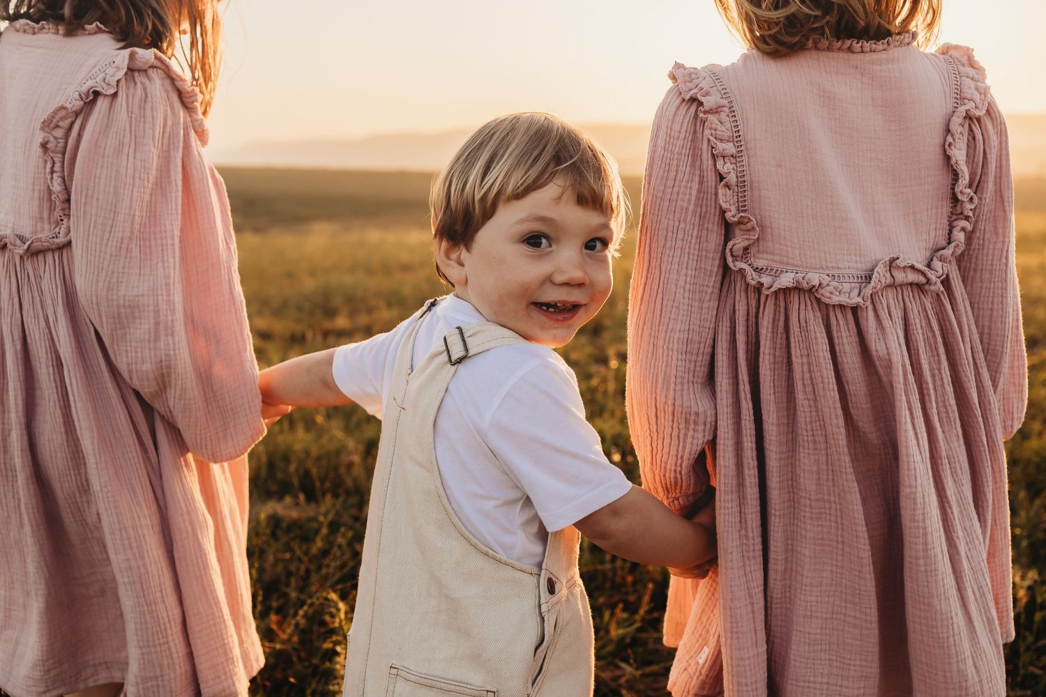 A group of people in soft pink and white dresses stand together in a sunlit field at golden hour.