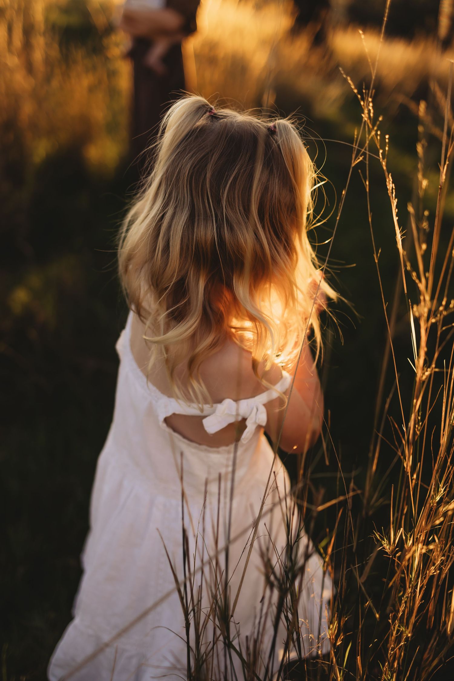 Person in white dress stands among tall golden grass at sunset with sunlight creating a dreamy backlit glow.