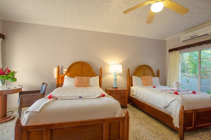 Twin bedroom with wooden beds, white linens, and ceiling fan in a tropical resort setting.