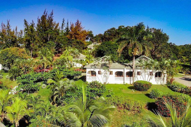 White Caribbean villa surrounded by lush tropical gardens and palm trees under bright blue sky.