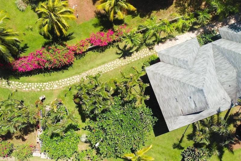 Aerial view of a tropical garden path with pink flowers leading to a white-roofed structure.