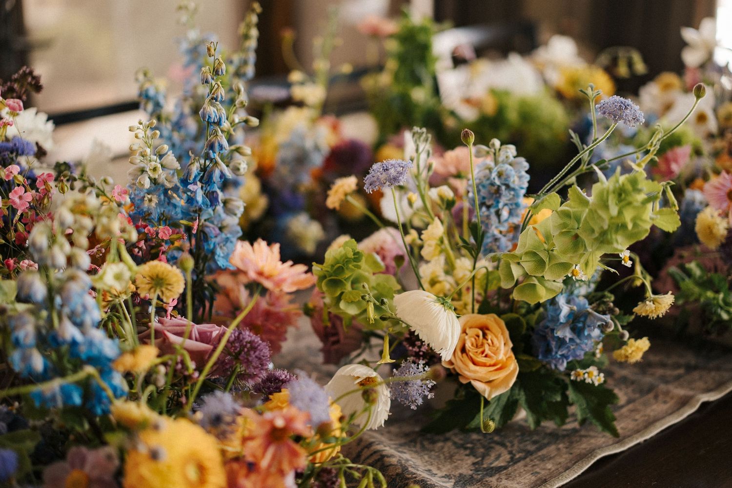 Colorful floral arrangements with peach roses and blue delphiniums decorate a rustic wooden table setting.