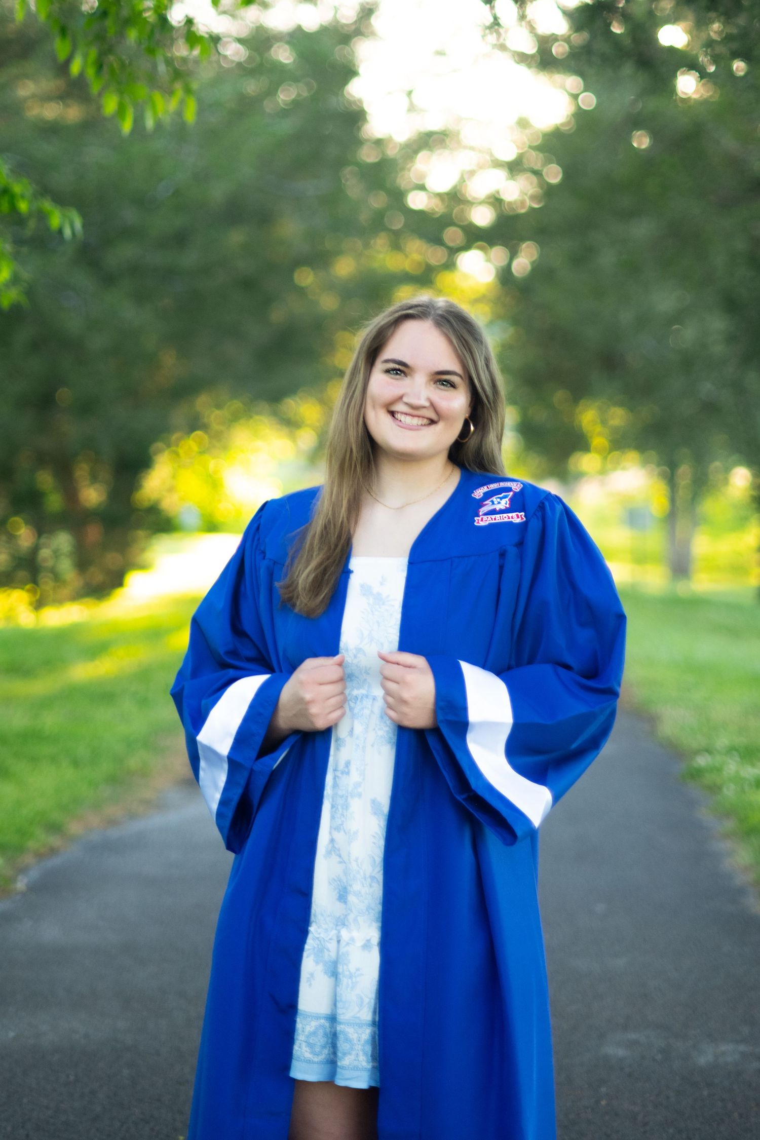 Graduate in blue cap and gown poses outdoors on a path with green trees and natural lighting in the background.