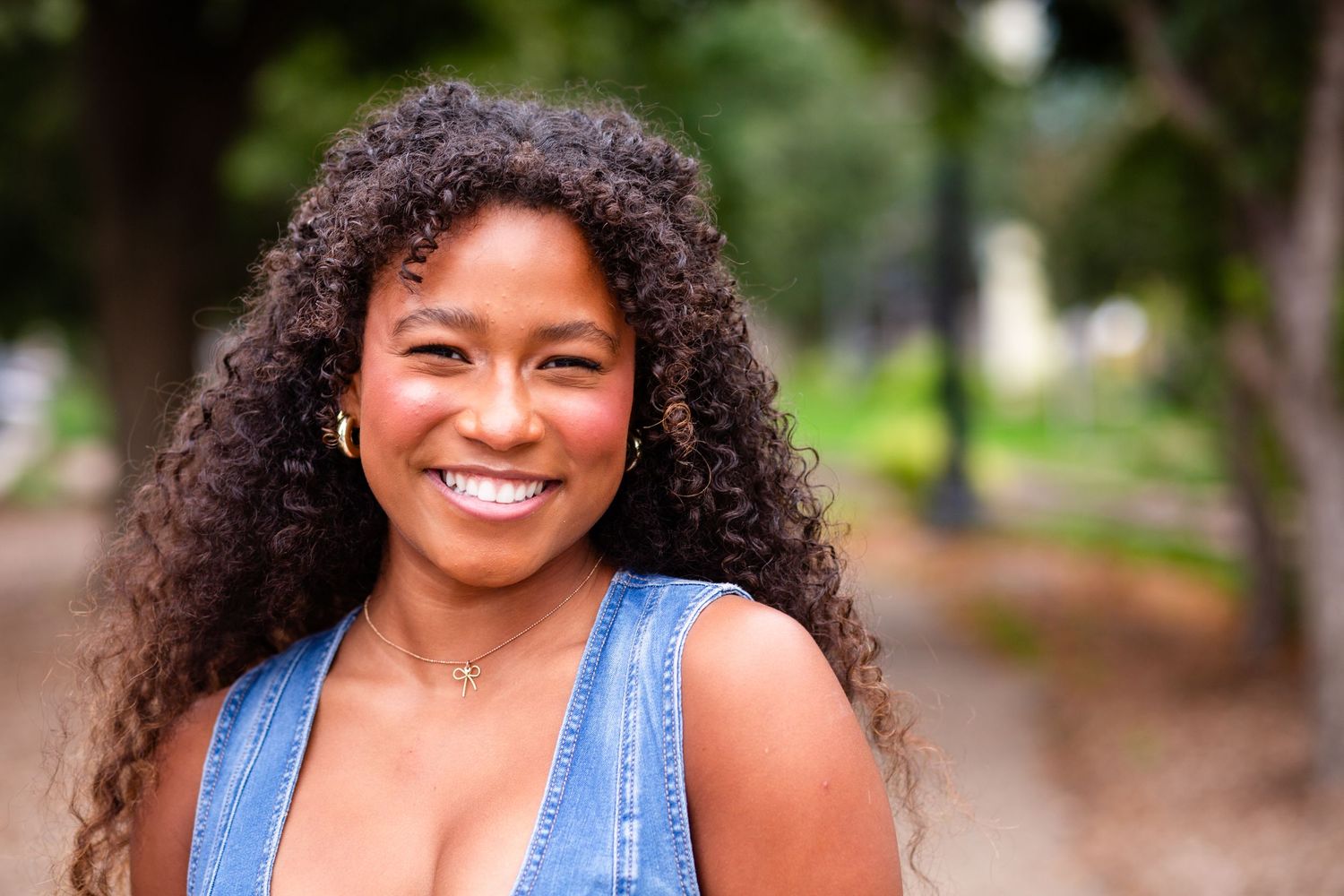 A high school senior girl with curly hair and a blue tank top smiles warmly at the camera in an outdoor setting downtown Cedar Rapids, IA.