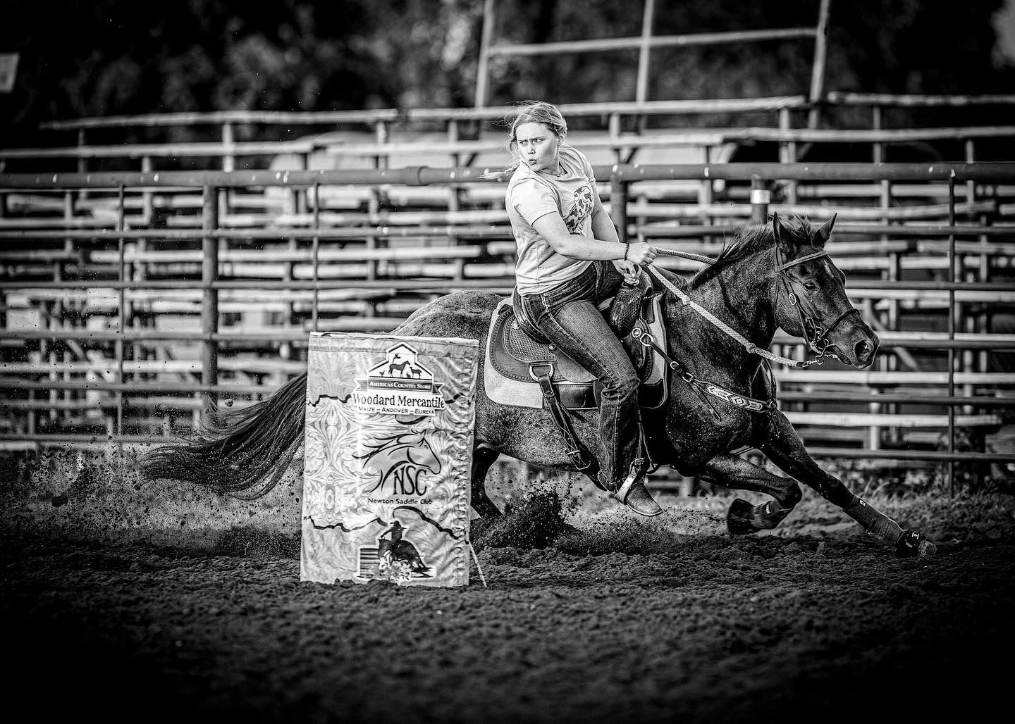 Black and white photo of a cowgirl riding a horse around a barrel during a rodeo competition.