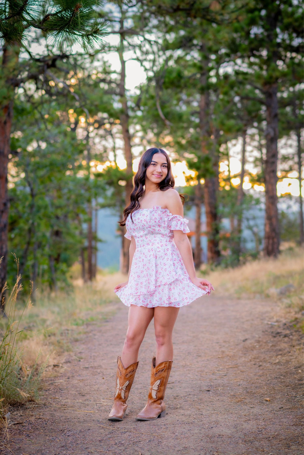 Child in floral dress and cowboy boots poses on forest trail at sunset.