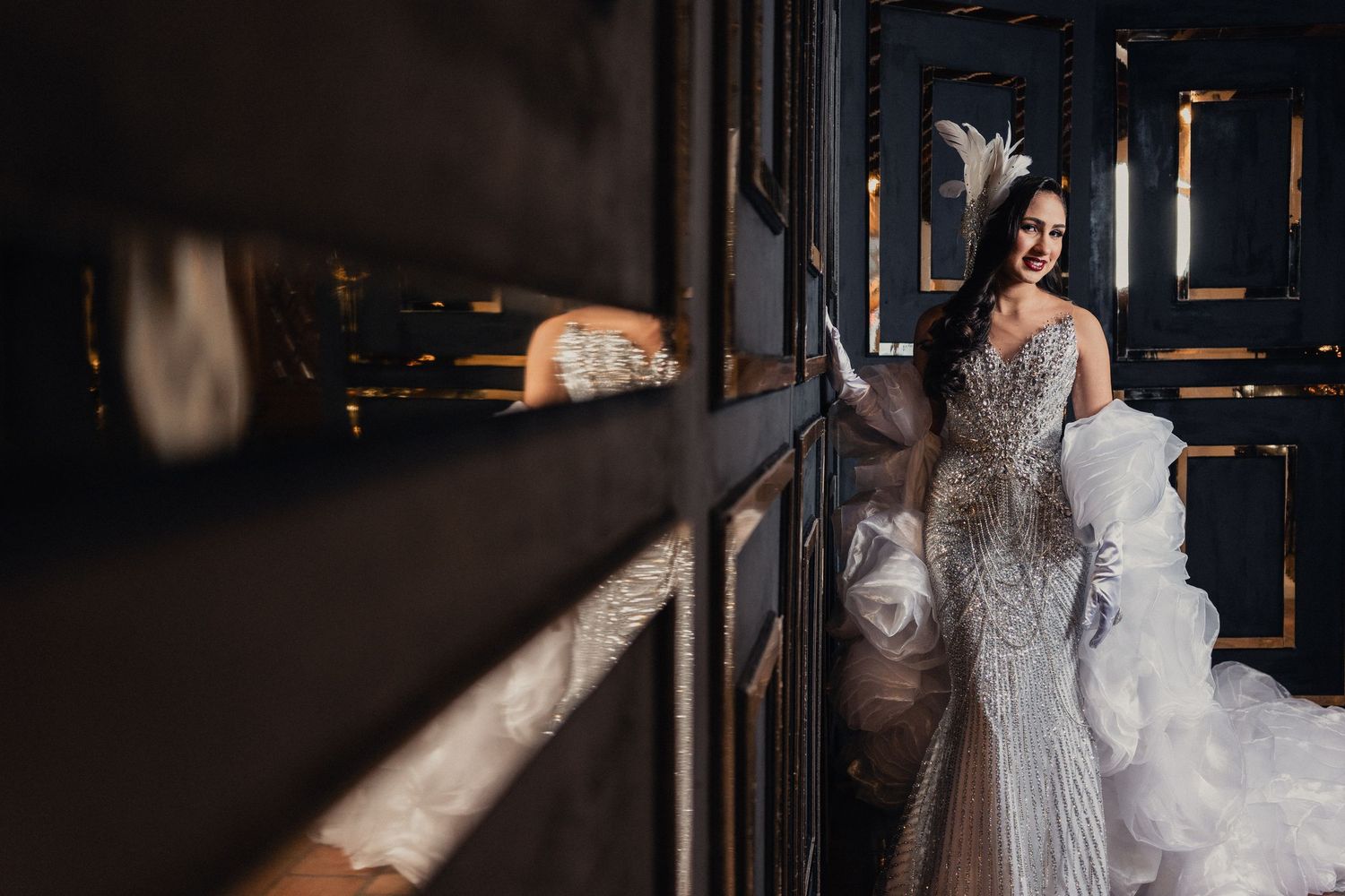 A bride in a white lace gown stands in dramatic lighting near a dark window creating a moody atmosphere.