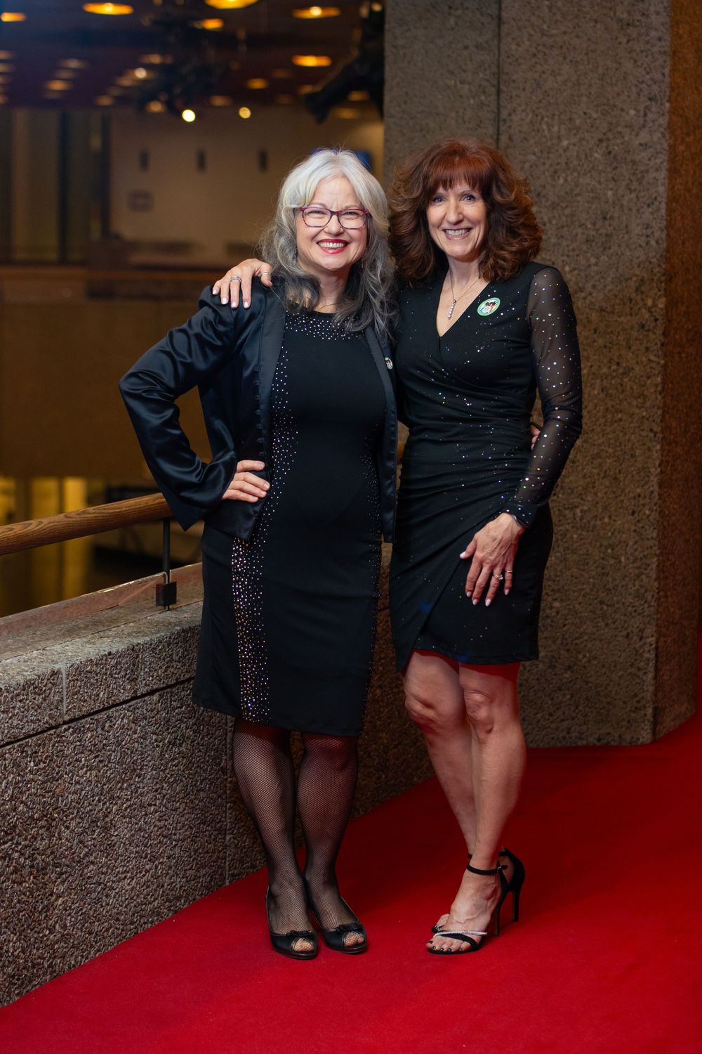 Two friends in black dresses pose together on a red carpet at an elegant evening event.
