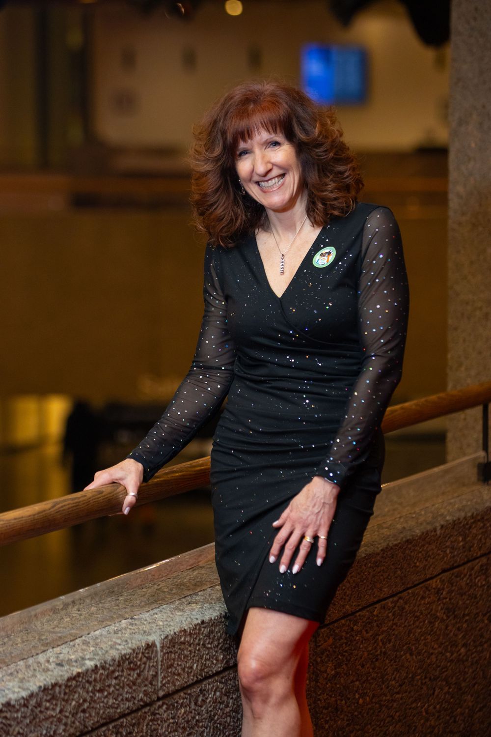 Woman in sparkly black dress poses elegantly on stairs in warm-lit venue.