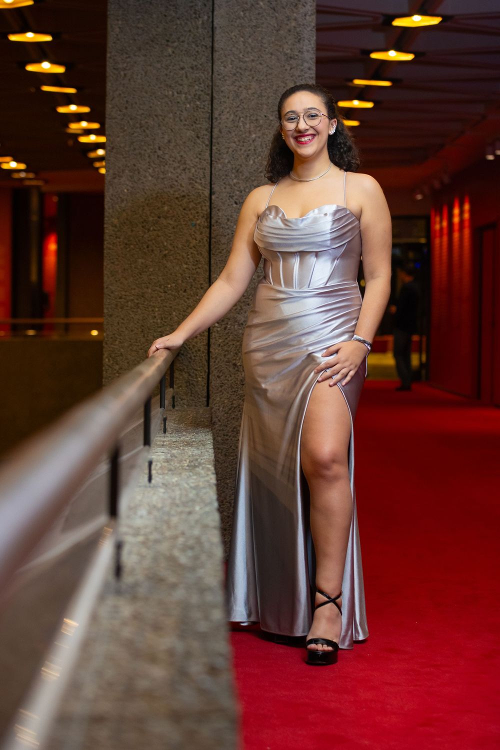 Person in silver dress poses gracefully along red-carpeted hallway with dramatic lighting.