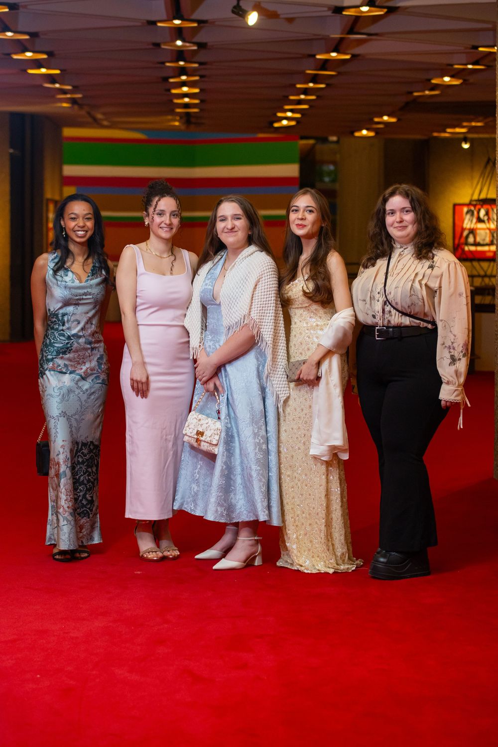 Five people in elegant dresses and formal attire pose together on a red carpet at an indoor event.