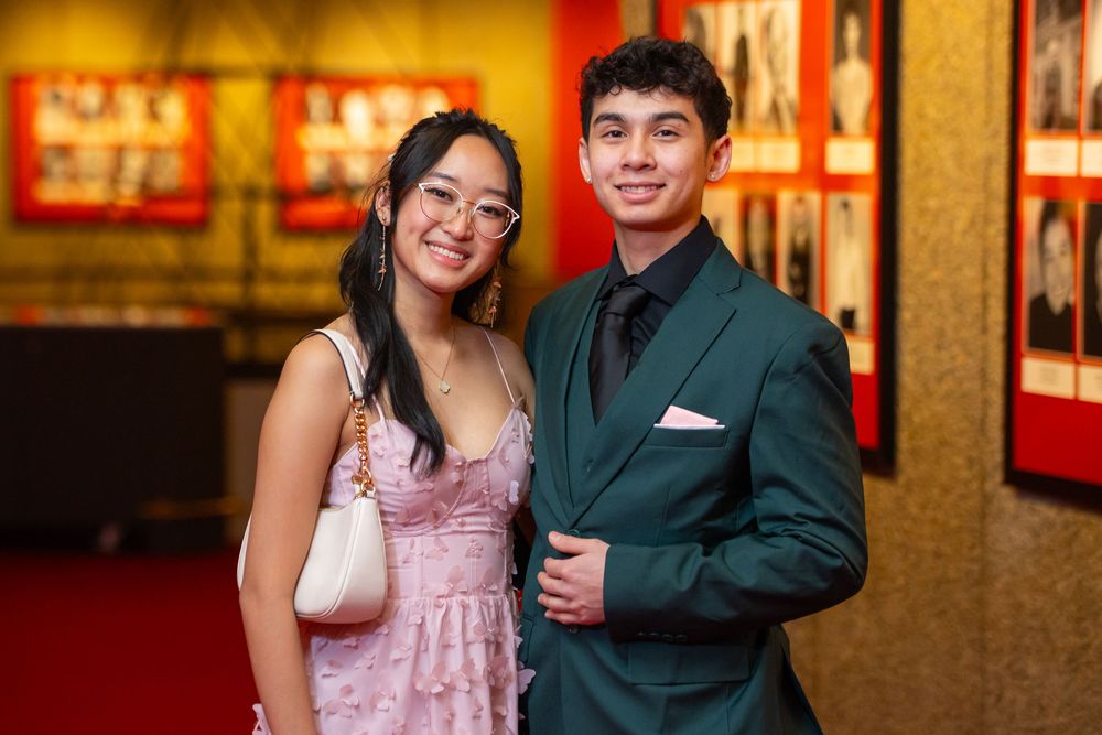 A couple in formal attire poses together at a venue with warm lighting and framed artwork on the walls.