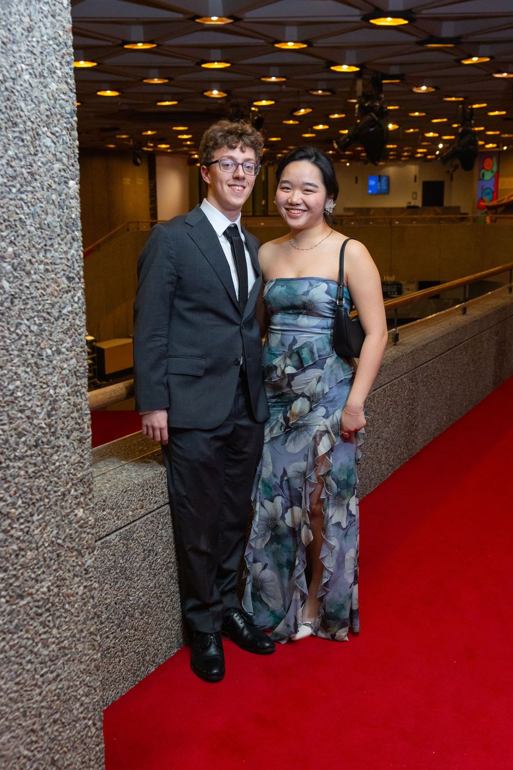 A couple poses on a red carpet, with one wearing a dark suit and the other in a floral dress.