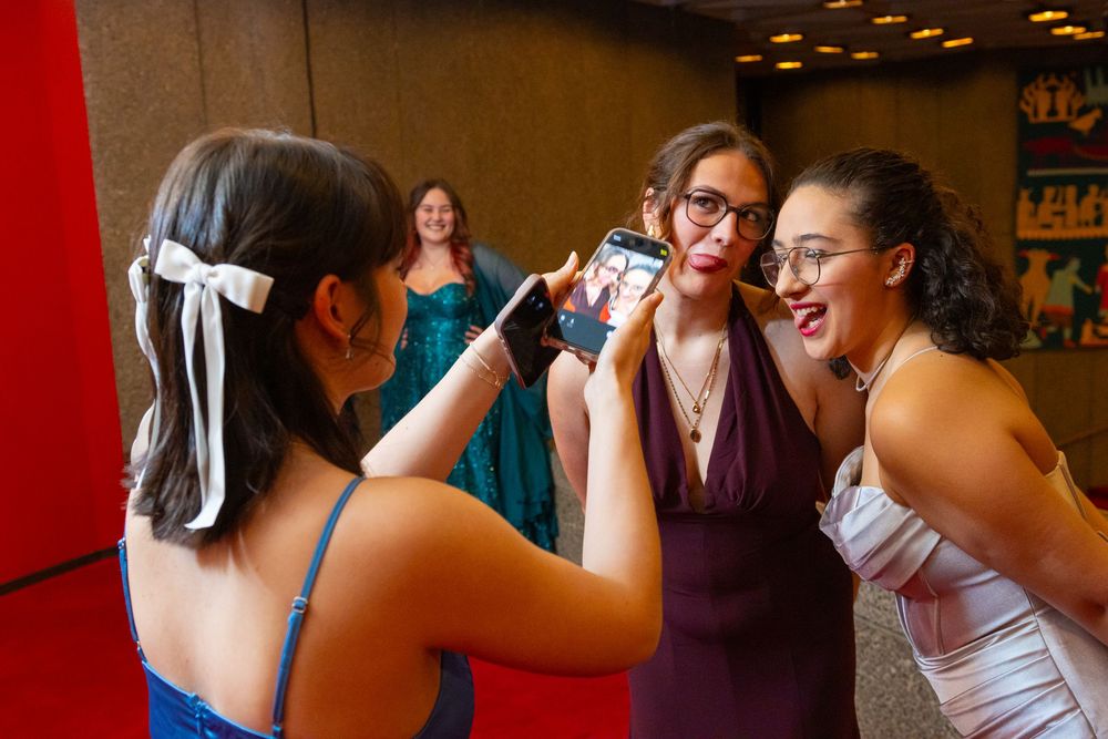 People laugh and take photos together at a formal event, with someone wearing a teal gown visible in the background.