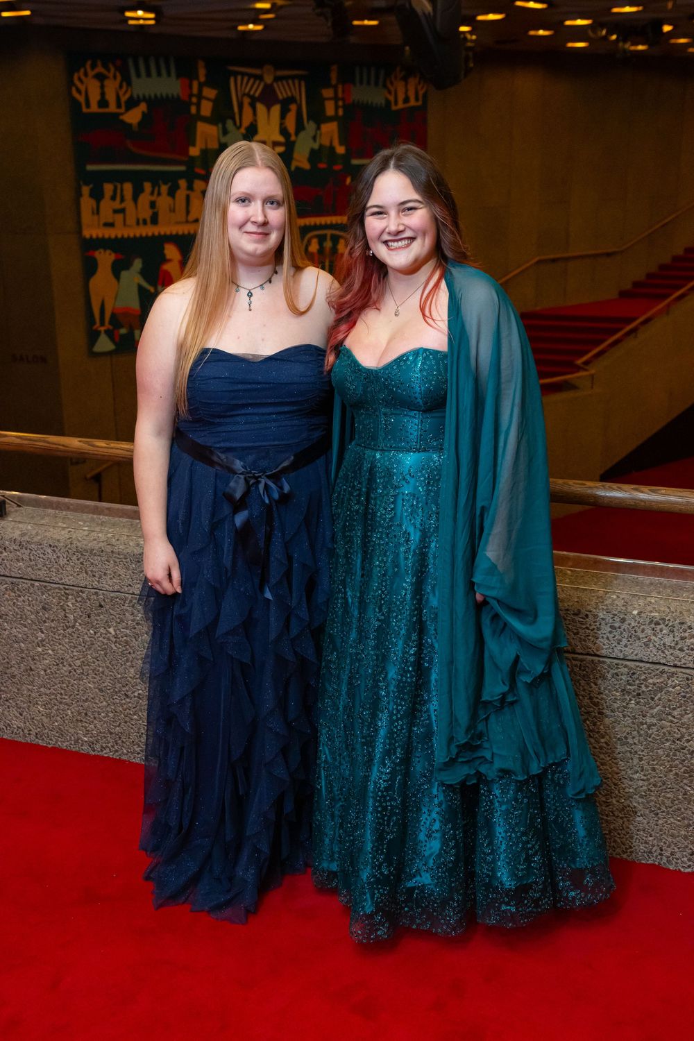 Two women pose together on a red carpet wearing elegant formal dresses in navy blue and sparkling teal.