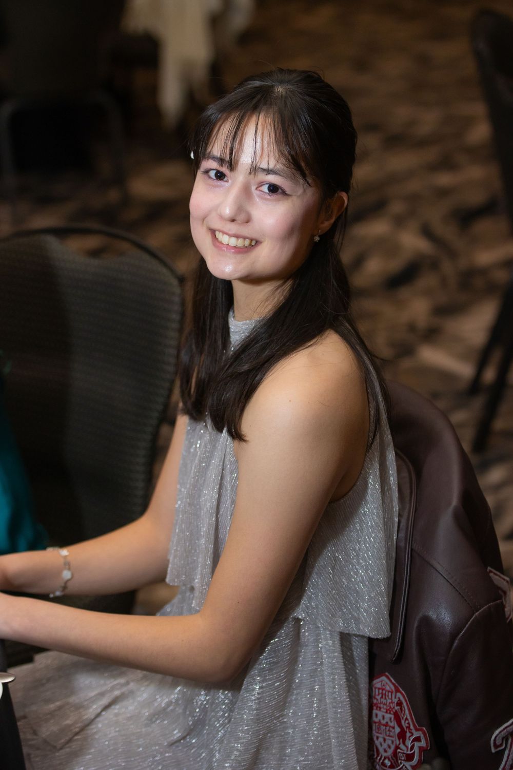 Young woman in a sparkly gray sleeveless dress smiles warmly while wearing a backpack in an indoor setting.