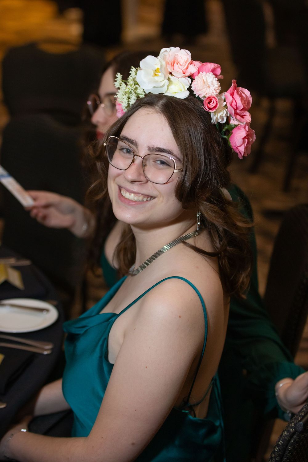 A smiling guest in a teal dress and glasses wears a pink and white floral crown at a formal event.