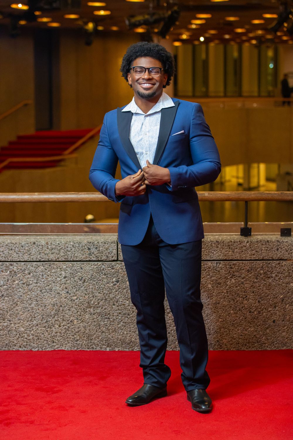Stylish man in blue suit and glasses poses confidently on red carpet at formal event.