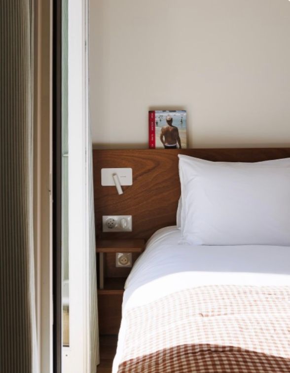 Modern hotel room interior with white bedding, wooden headboard with built-in power outlets, and neutral decor.